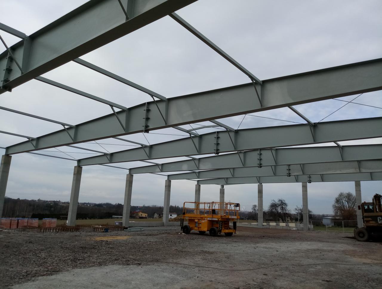 Stalowa konstrukcja szkieletowa budynku w trakcie budowy, widoczna żółta platforma robocza i stos cegieł w tle.