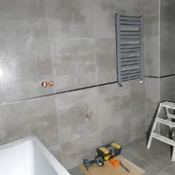 Szare płytki ścienne w łazience, zamontowany grzejnik drabinkowy, widoczne przygotowania do montażu armatury, biała wanna w rogu, narzędzia i skrzynka narzędziowa na podłodze.