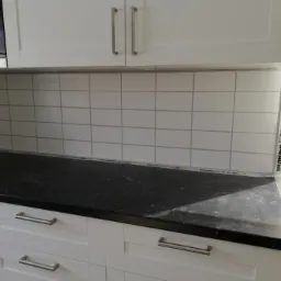 Białe, kwadratowe płytki ułożone na ścianie nad blatem kuchennym, zabezpieczone listwami ochronnymi K-Rauta podczas prac wykończeniowych w kuchni.