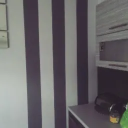 Ściana z trzema pionowymi pasami w kolorze szarym i białym, obok szare szafki kuchenne z metalowymi uchwytami, widoczne akcesoria kuchenne na blacie.