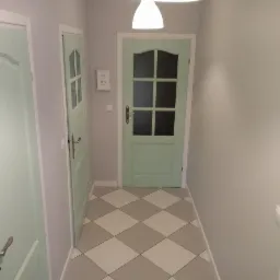 Korytarz z dwiema pomalowanymi na pastelowo zielono drzwiami, podłoga wyłożona szaro-białą szachownicą z płytek, ściany w odcieniach szarości, widoczna lampa sufitowa w kształcie stożka.