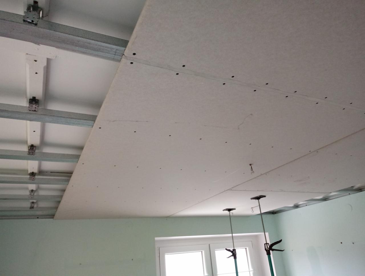 Montaż sufitu podwieszanego z płyt gipsowo-kartonowych na metalowej konstrukcji, widoczne podpory teleskopowe i ślady szpachlowania na łączeniach płyt.