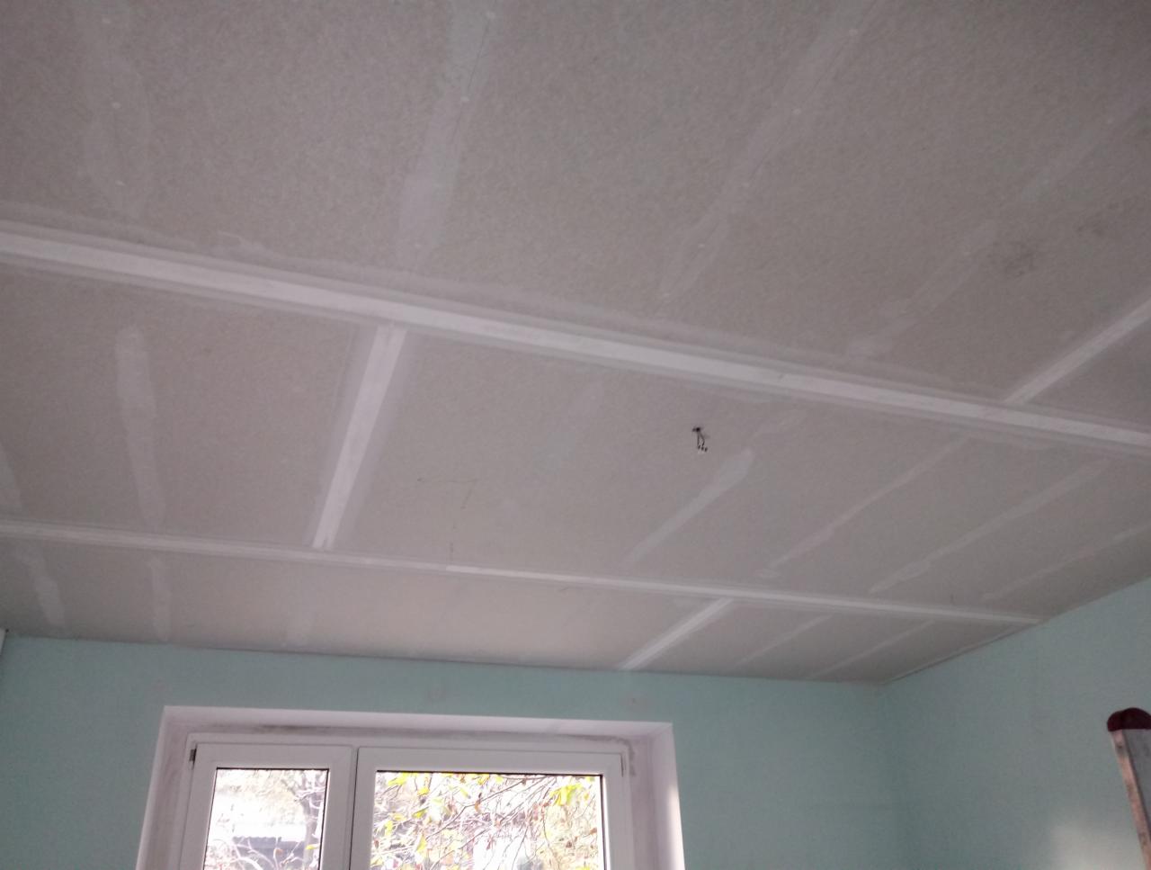 Sufit z płyt kartonowo-gipsowych w trakcie montażu, widoczne łączenia płyt, ściany pomalowane na jasny niebieski kolor, okno w tle.