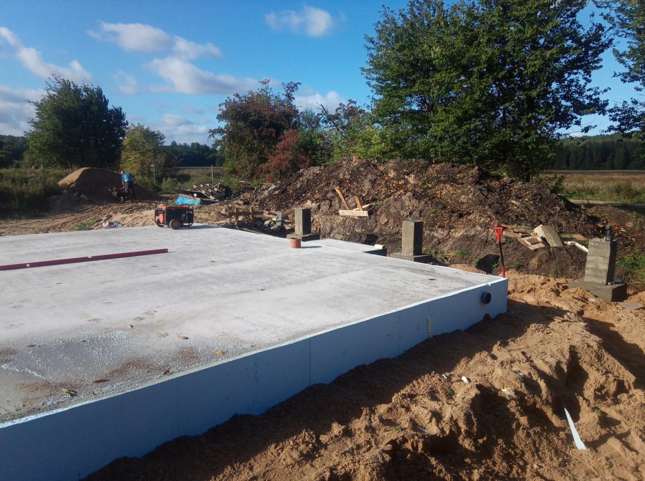 Wylany fundament z izolacją termiczną, widoczne słupki betonowe i stos ziemi w tle, poziomica na fundamencie, teren budowy w słoneczny dzień.