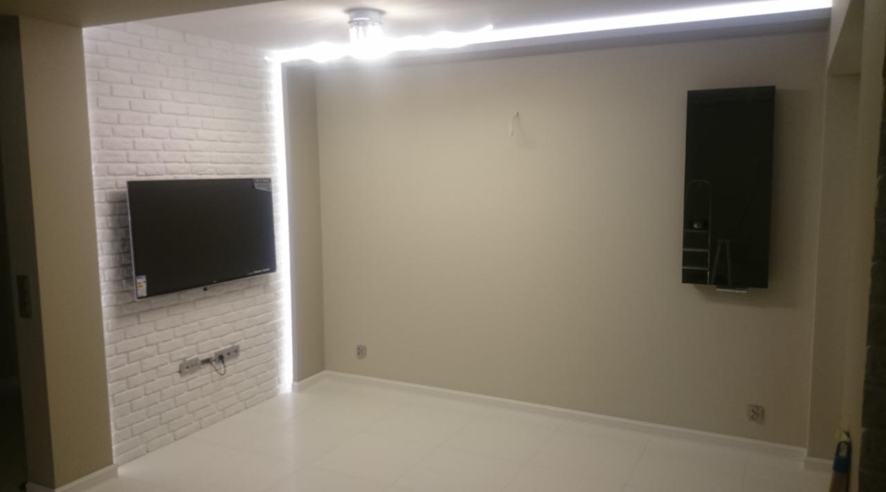 Salon po remoncie z białą cegłą dekoracyjną na ścianie, na której wisi telewizor. Na suficie widoczne oświetlenie LED. Na ścianie po prawej stronie wisi czarna szafka.
