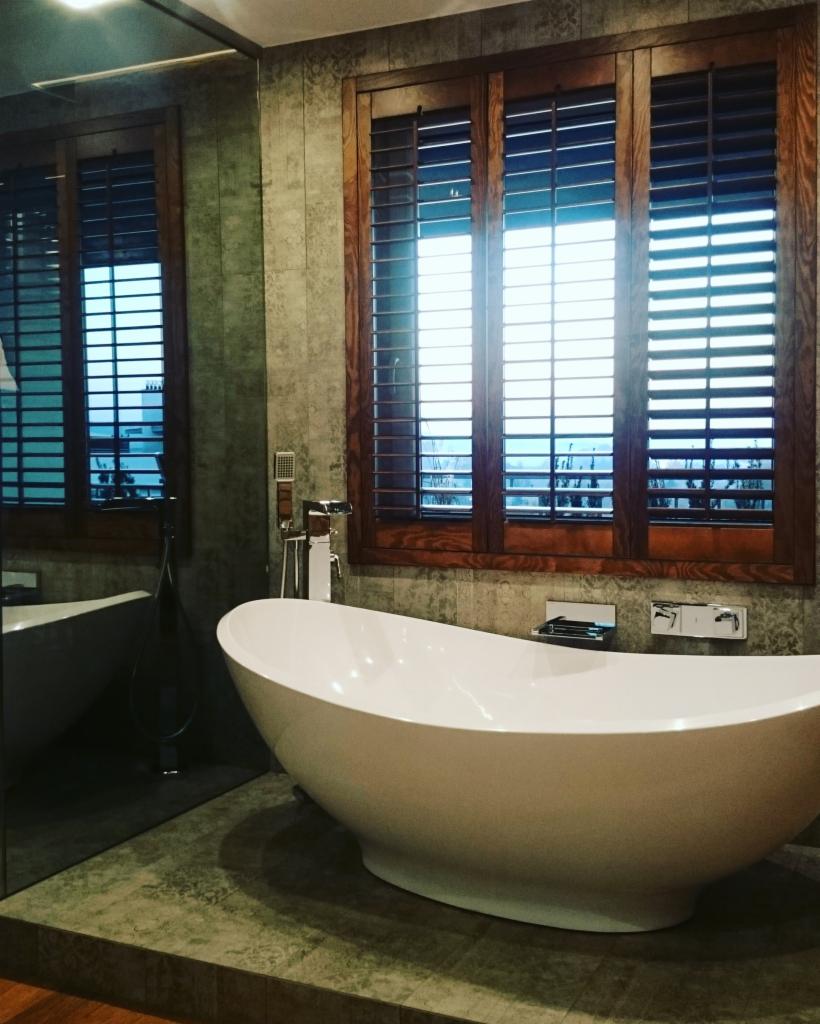 Nowoczesna łazienka z wolnostojącą białą wanną, drewnianymi żaluzjami i szarą mozaiką na ścianach, odbicie w dużym lustrze.