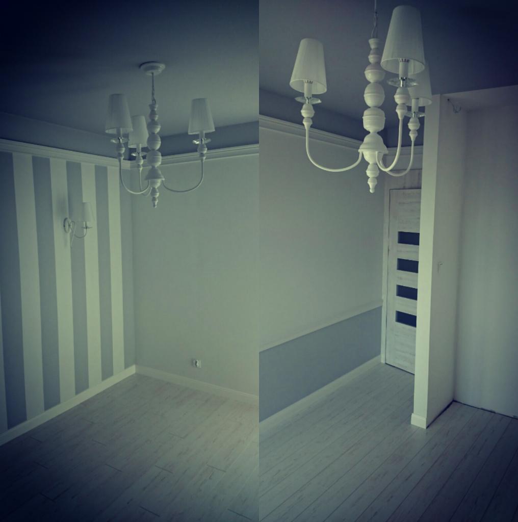 Pusta, jasna przestrzeń z pomalowanymi w pionowe pasy ścianami, ozdobnym żyrandolem z białymi abażurami i podłogą imitującą drewno, widok na drzwi z przeszkleniami.