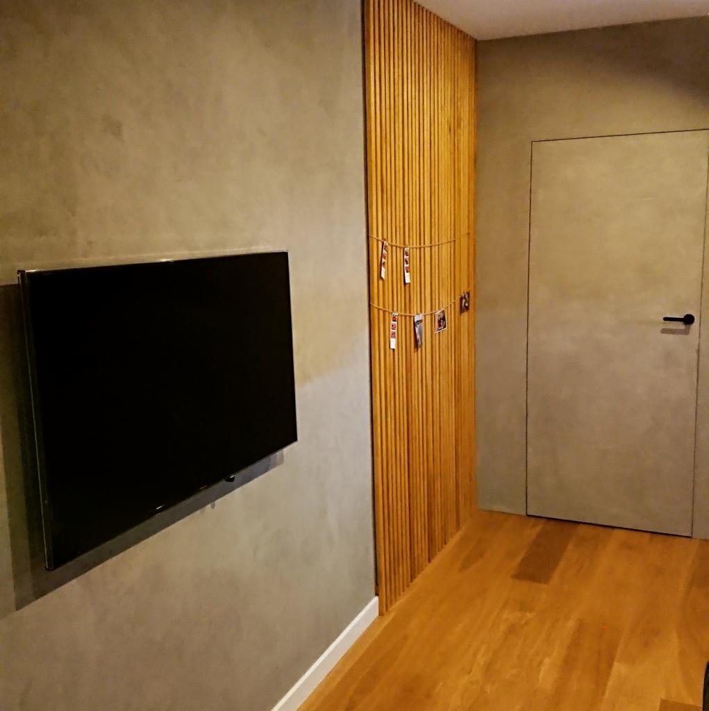 Szara ściana z telewizorem, pionowe drewniane panele z dekoracyjną girlandą ze zdjęć i dopasowane kolorystycznie drzwi w nowoczesnym wnętrzu.