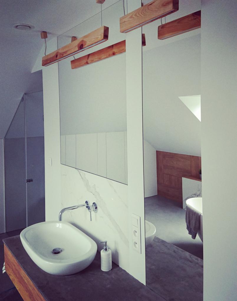 Nowoczesna łazienka z owalną umywalką nablatową, marmurową ścianą, drewnianymi elementami oświetlenia i widokiem na wannę wolnostojącą w lustrze.