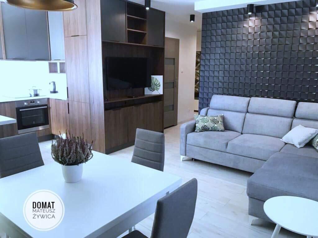 Nowoczesne wnętrze: szara sofa narożna z poduszkami na tle ściany z panelami 3D, biały stół z dekoracją kwiatową, widoczny fragment kuchni z drewnianymi szafkami i piekarnikiem.