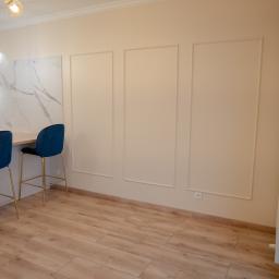 KWW Marcin Rzepniewski - Jasne wnętrze po remoncie: panelowa ściana, drewnopodobna podłoga, marmurowa ściana w kuchni, dwa granatowe hokery z złotymi nogami i designerskie lampy.