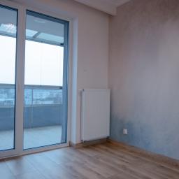 KWW Marcin Rzepniewski - Pusty pokój po odświeżeniu z panelami podłogowymi, grzejnikiem, pomalowaną ścianą z efektem ombre i dużym oknem balkonowym z widokiem na miasto.