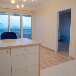 KWW Marcin Rzepniewski - Jasne wnętrze mieszkania z drewnianą podłogą, widokiem na miasto przez okno i meblami w odcieniach beżu, oświetlone designerską lampą.