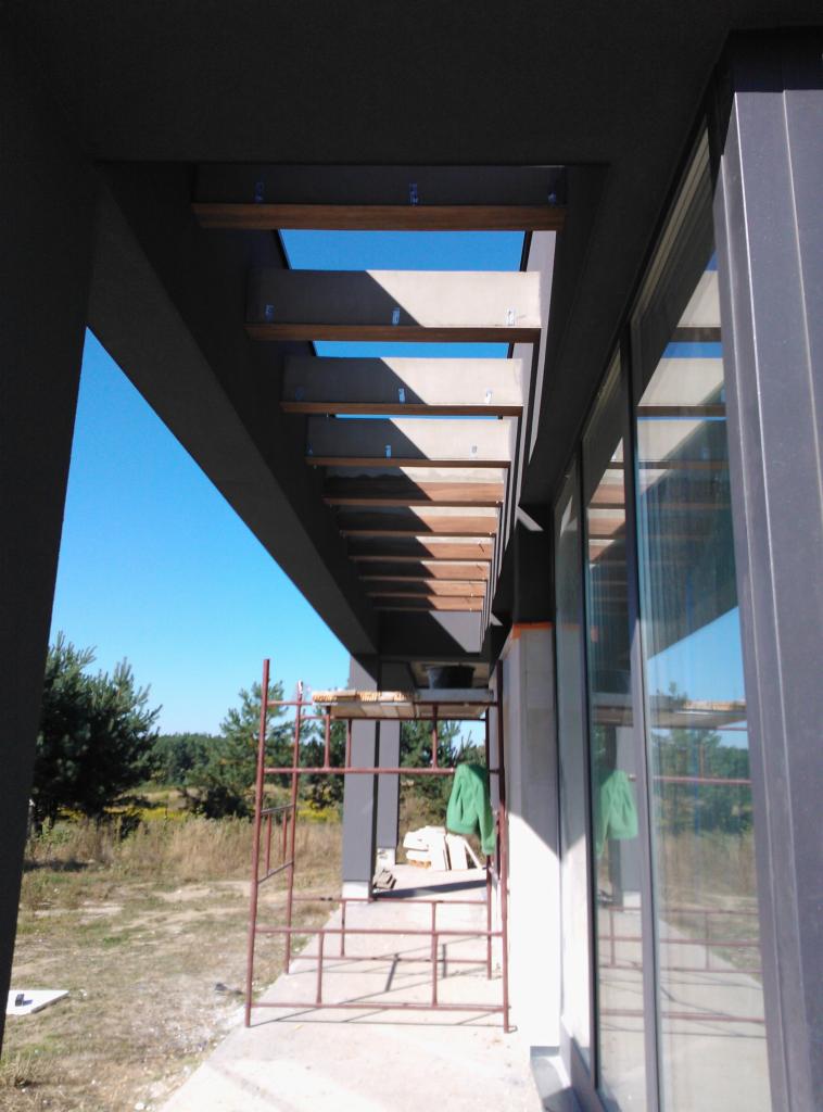 Montaż drewnianych belek pod zadaszeniem tarasu, widoczne rusztowanie i okno z odbiciem krajobrazu.