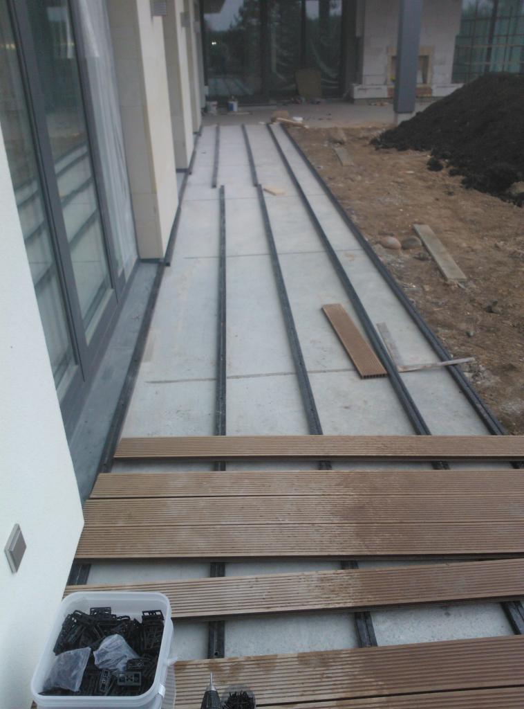 Montaż deski kompozytowej na tarasie: widoczne legary, częściowo ułożone deski, pojemnik z klipsami montażowymi i śrubokręt.