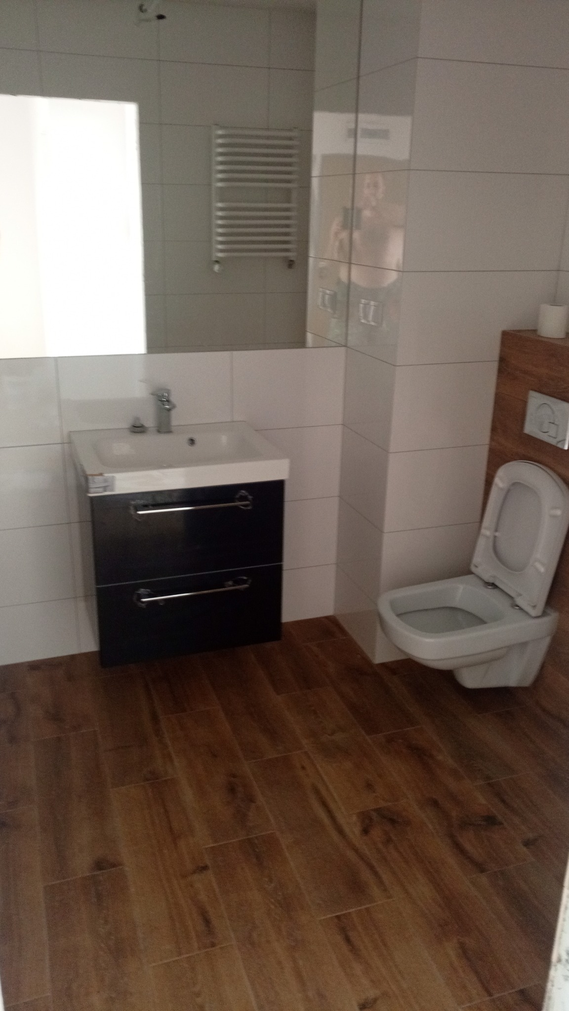 Nowoczesna łazienka z drewnopodobną podłogą, białymi płytkami ściennymi, wiszącą szafką z umywalką w kolorze czarnym oraz toaletą. W lustrze odbija się osoba robiąca zdjęcie.