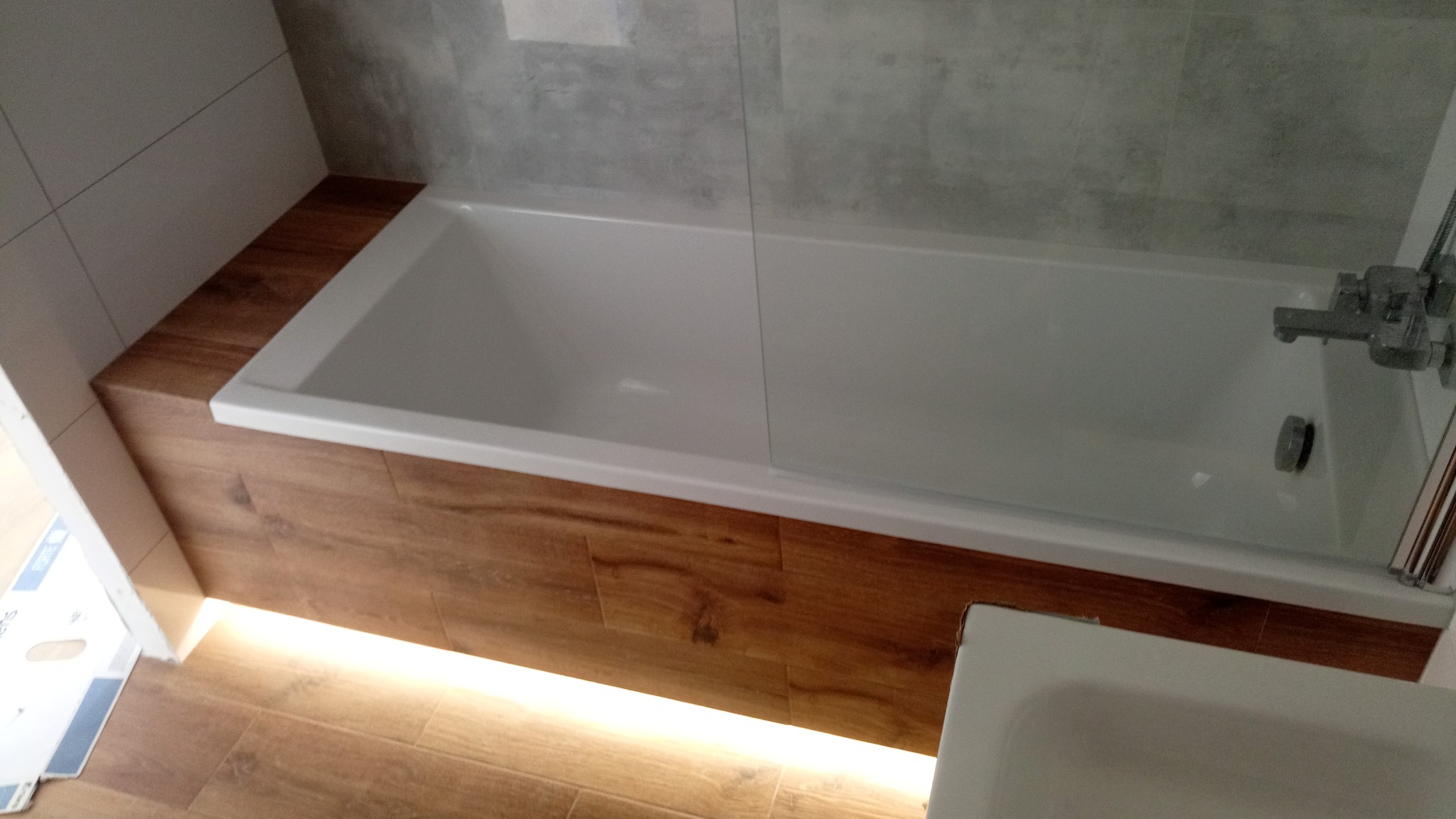 Nowoczesna biała wanna obudowana drewnopodobnymi panelami z subtelnym podświetleniem LED u dołu, szare płytki na ścianie w tle.