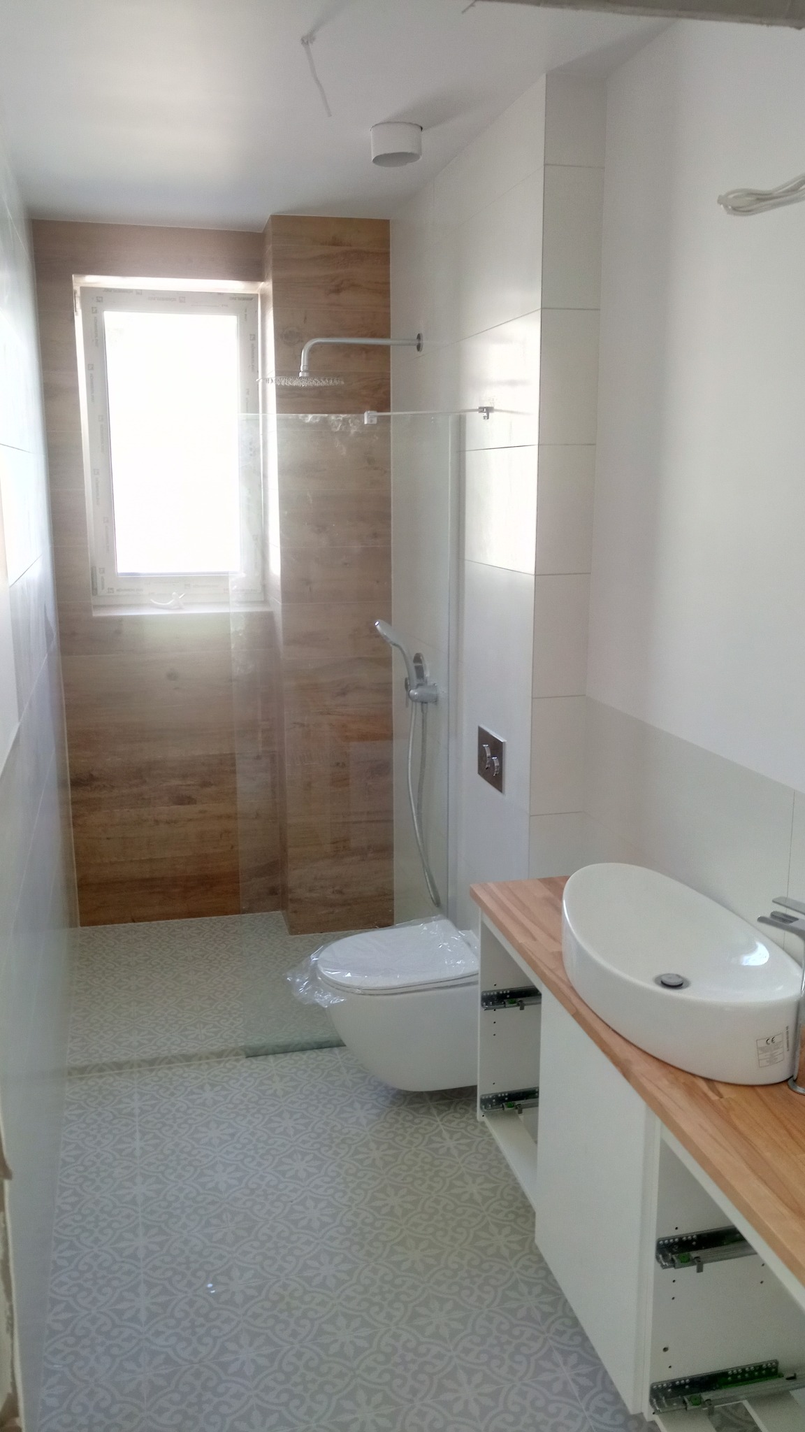 Nowoczesna łazienka z kabiną prysznicową typu walk-in, umywalką nablatową na drewnianym blacie, toaletą w folii ochronnej i wzorzystą podłogą.