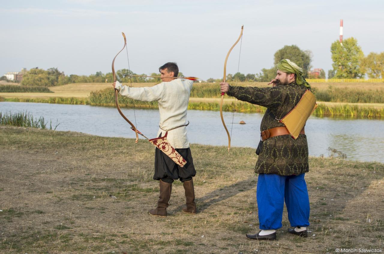 Dwóch mężczyzn ubranych w stroje historyczne, naciągających łuki na tle jeziora i krajobrazu, z widocznym kołczanem i turbanem.