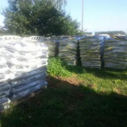 Ułożone na paletach, owinięte folią worki z opałem na trawiastym terenie, widoczne drzewa i fragment budynku w tle.