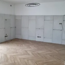 Wnętrze w trakcie remontu: ułożona podłoga z paneli w jodełkę, ściany wyłożone płytami kartonowo-gipsowymi z widocznymi otworami na instalacje elektryczne i gniazdka, nowoczesna lampa sufitowa.