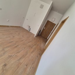 Remonty I Wykończenia Wnętrz Dominika Szczęsna - Jasne wnętrze po remoncie: drewnopodobne panele, białe ściany, drzwi wejściowe z brązowego drewna. Widoczne gniazdka i elementy instalacji.