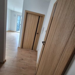 Remonty I Wykończenia Wnętrz Dominika Szczęsna - Nowe drewniane drzwi wewnętrzne z czarnymi akcentami, widok na parkiet i odległe okno w wyremontowanym mieszkaniu. Minimalistyczny design.