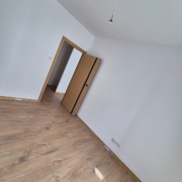 Remonty I Wykończenia Wnętrz Dominika Szczęsna - Jasne panele podłogowe w pokoju z białymi ścianami i drewnianymi drzwiami z czarnymi akcentami. Widoczne gniazdka elektryczne i wystający kabel z sufitu.