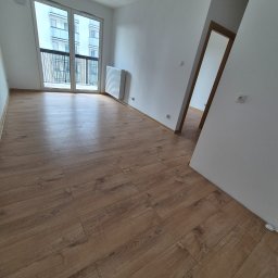 Remonty I Wykończenia Wnętrz Dominika Szczęsna - Jasne panele podłogowe w nowym, pustym pokoju z balkonem i grzejnikiem. Widok na otwarte drzwi do sąsiedniego pomieszczenia. Minimalistyczny design.