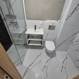 Biała łazienka w stylu marmuru z kabiną prysznicową, umywalką z szafką i toaletą. Surowy, betonowy sufit kontrastuje z eleganckimi płytkami.