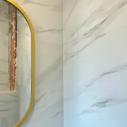 Białe płytki imitujące marmur na ścianie łazienki, z widoczną rurą obudowaną pianką montażową i fragmentem złotego lustra.