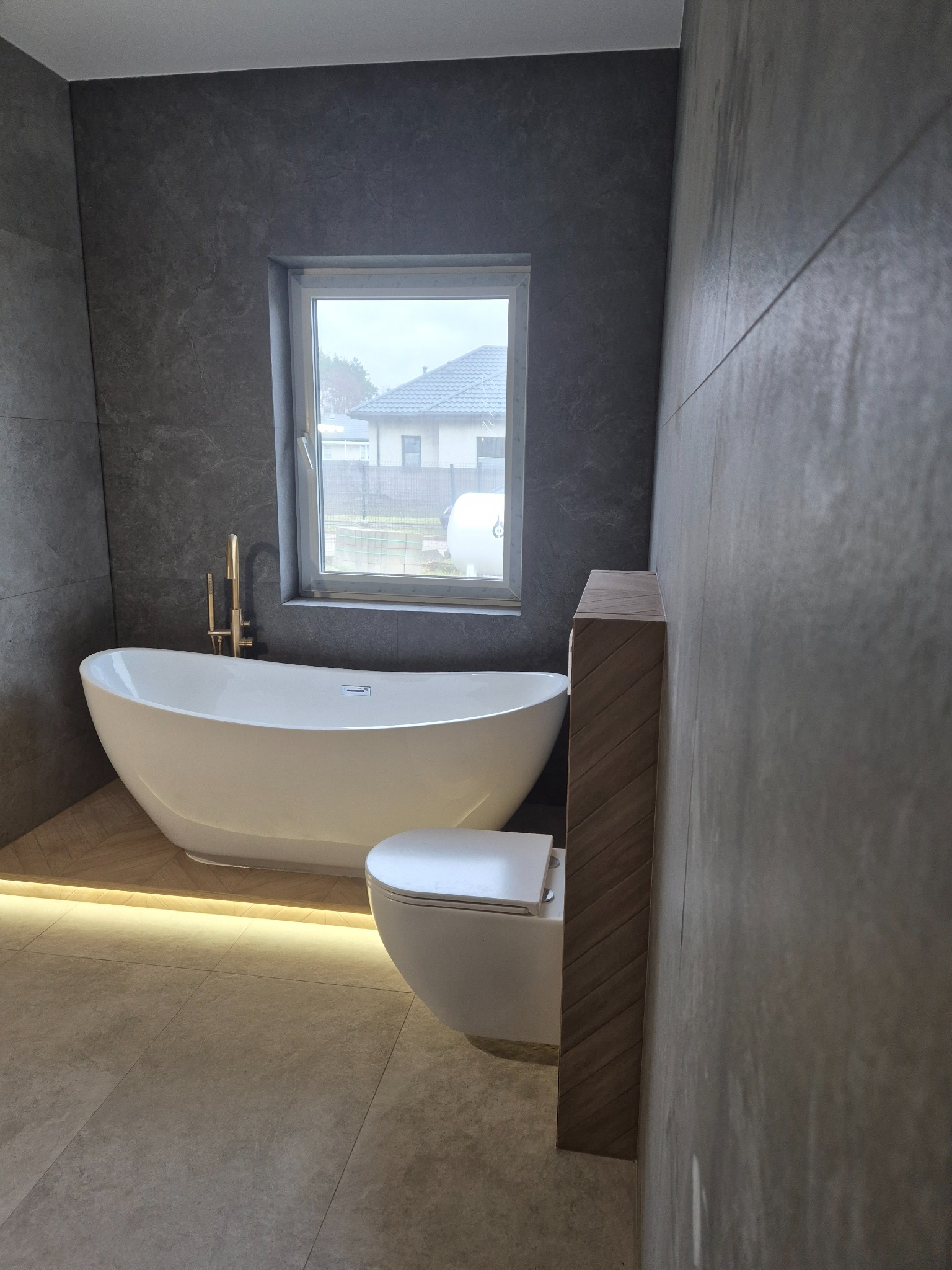 Nowoczesna łazienka z wolnostojącą wanną, złotą armaturą i podświetleniem LED pod wanną, widok na dom przez okno.