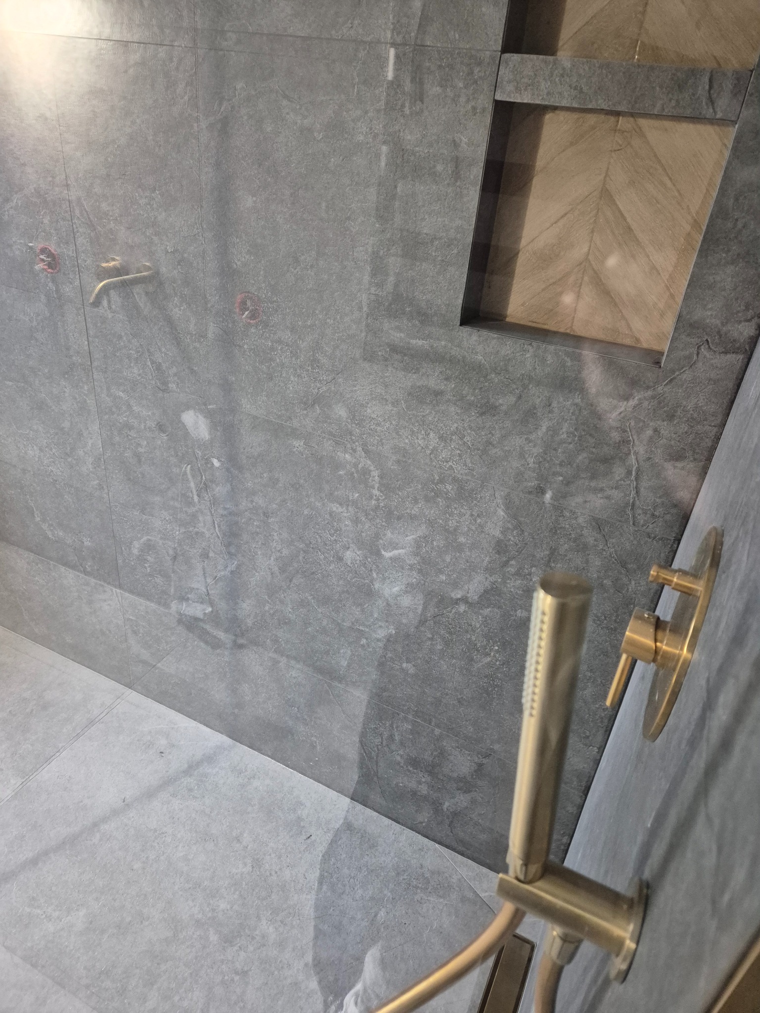 Szara, marmurkowa łazienka z wnęką wyłożoną drewnem w jodełkę i złotą armaturą prysznicową.