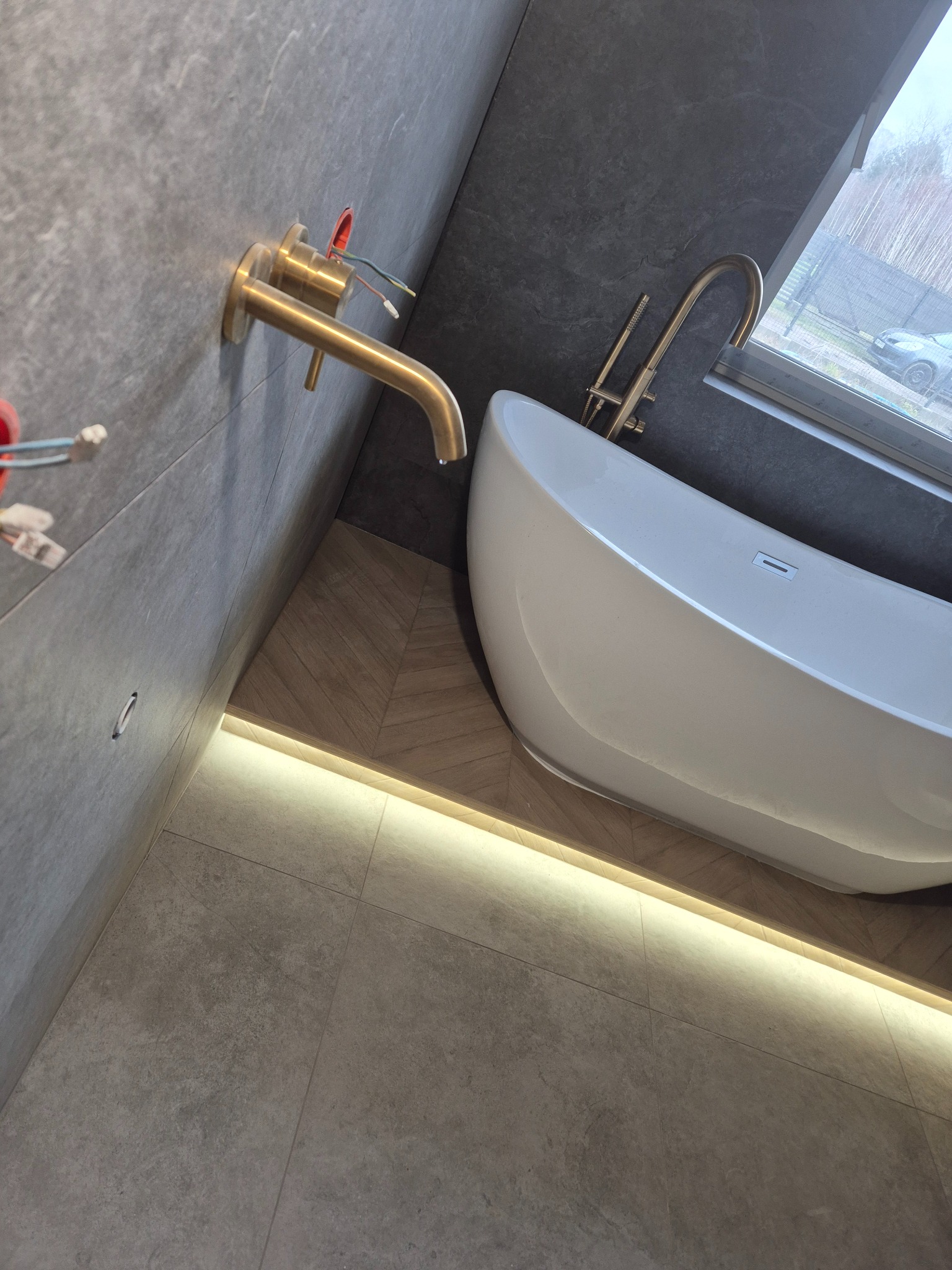 Nowoczesna łazienka z wolnostojącą białą wanną, złotą armaturą i podświetleniem LED wzdłuż podłogi i ściany.