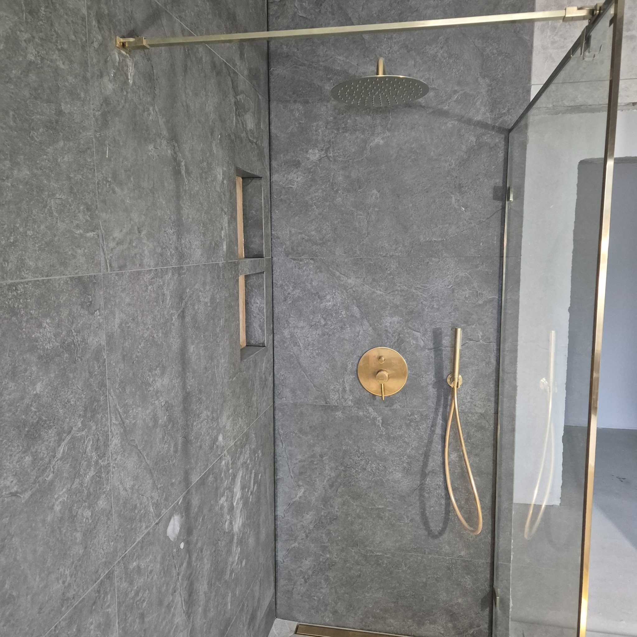 Szara łazienka z deszczownicą i złotą armaturą, widoczny fragment kabiny prysznicowej z szklaną ścianą i złotymi profilami, w ścianie wnęki wykończone złotym obramowaniem.