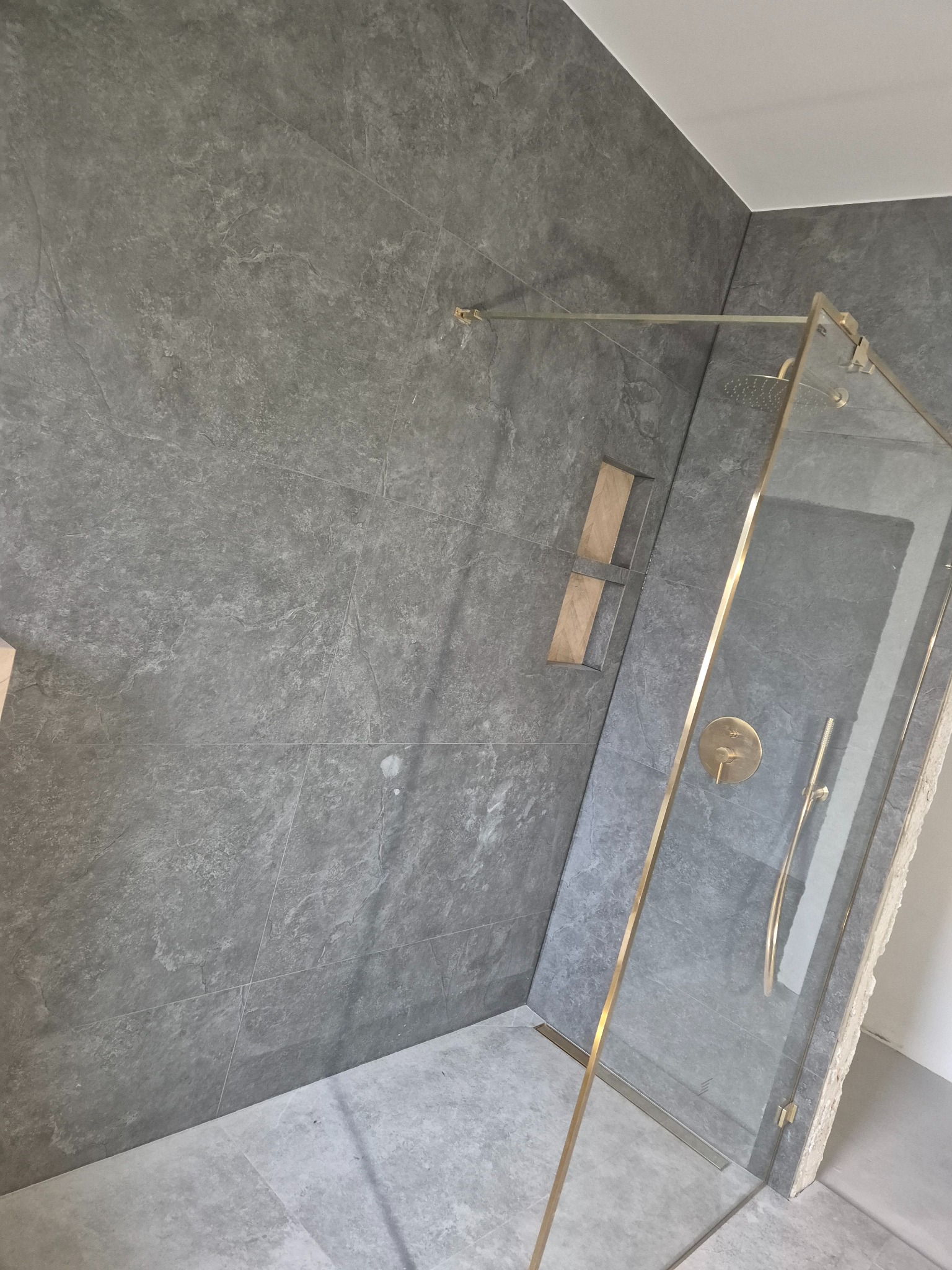 Szara łazienka z kabiną prysznicową ze złotymi elementami, wbudowana półka w ścianie wyłożonej płytkami imitującymi kamień.