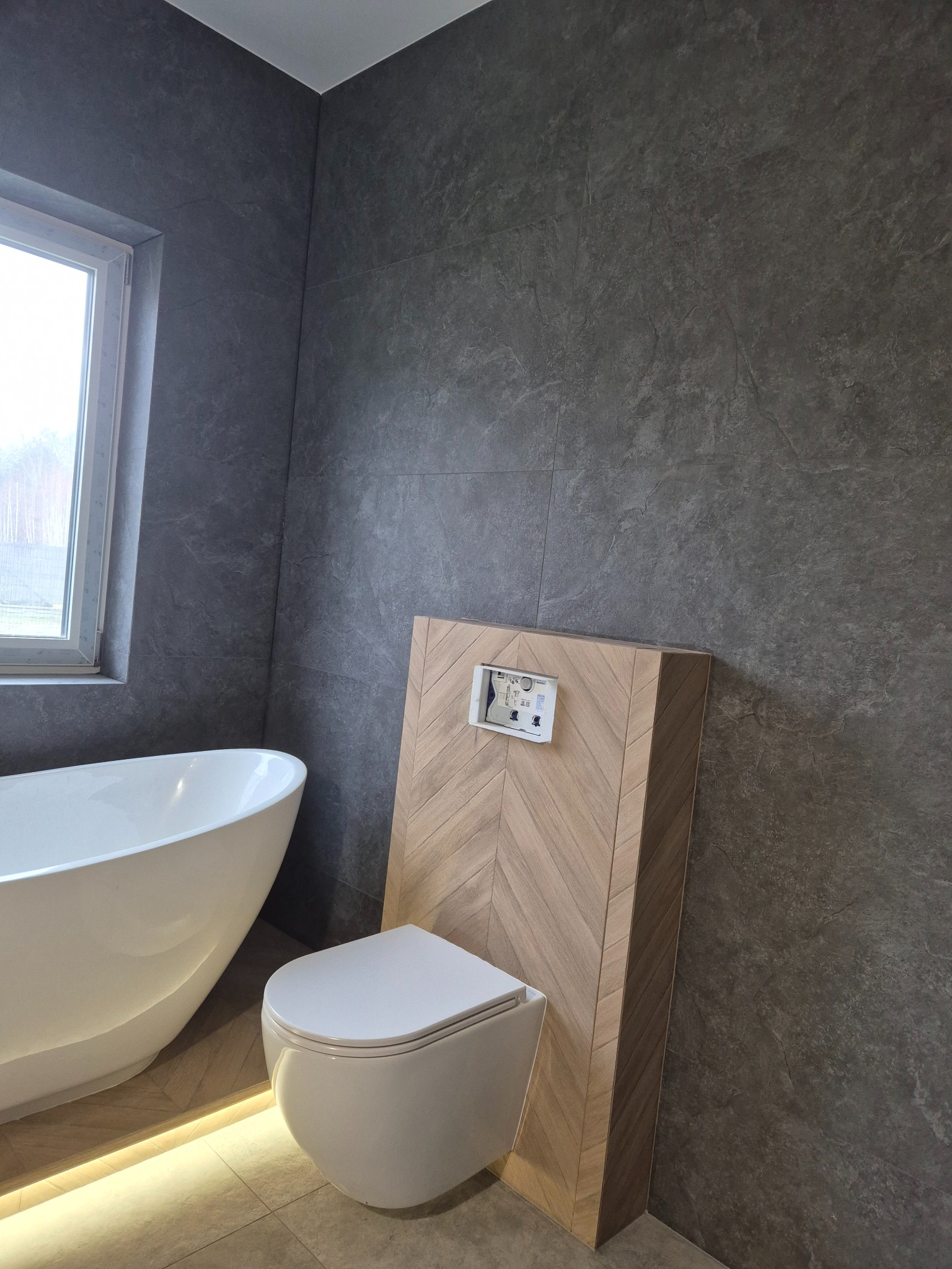 Nowoczesna łazienka z wolnostojącą wanną, podświetleniem LED i toaletą wiszącą zabudowaną drewnianą mozaiką w jodełkę, szare płytki imitujące kamień na ścianach.