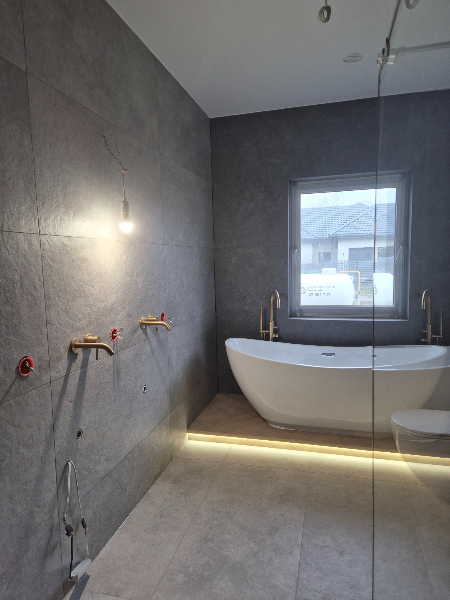 Szare płytki w łazience w Grodzisku Mazowieckim, widoczne przygotowane przyłącza do baterii, wanna wolnostojąca i podświetlenie podłogowe w trakcie montażu.