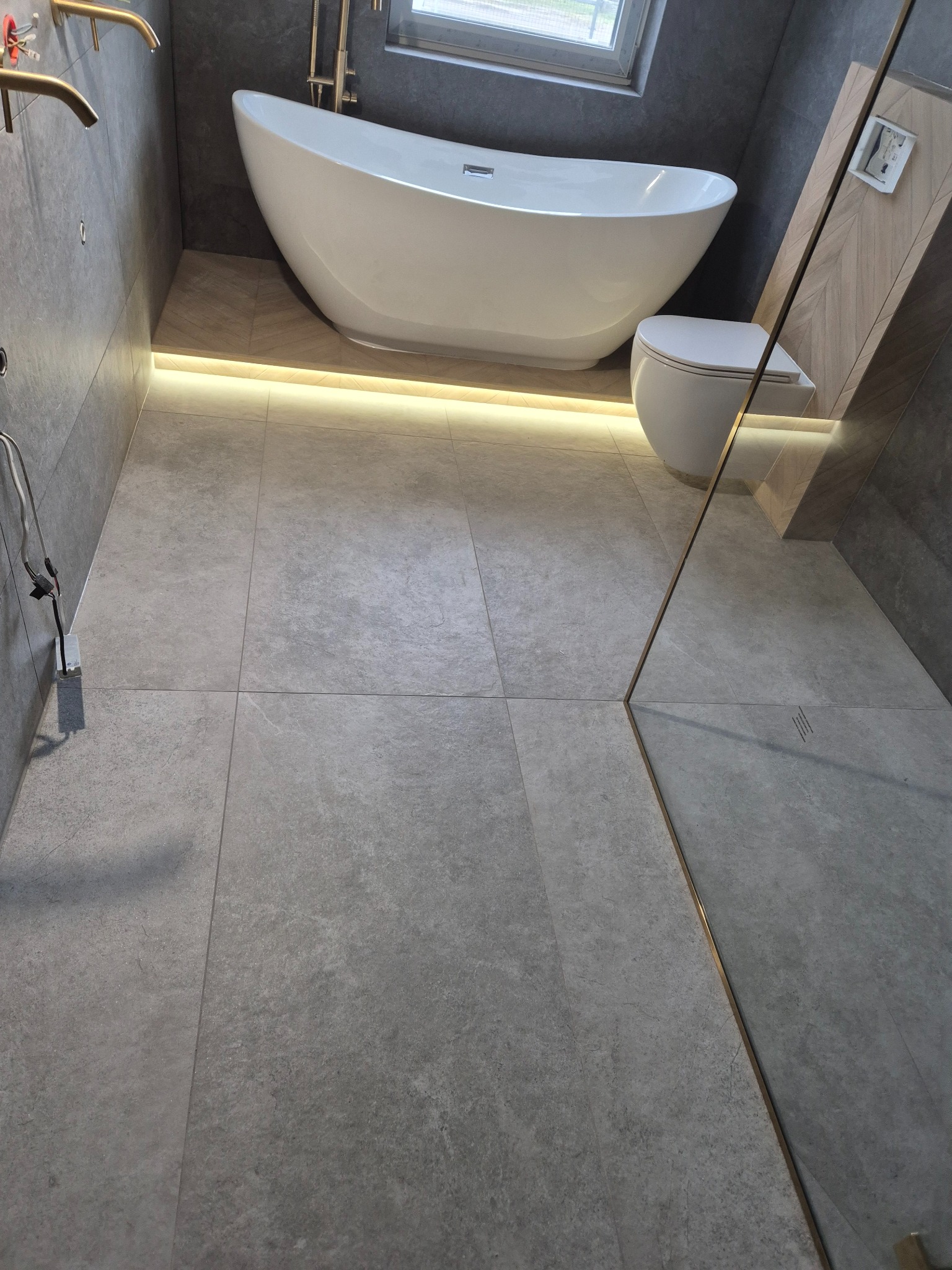 Nowoczesna łazienka z wolnostojącą wanną, podświetleniem LED pod wanną i szarą płytką podłogową o matowym wykończeniu, widok na kabinę prysznicową z elementami w kolorze złotym.