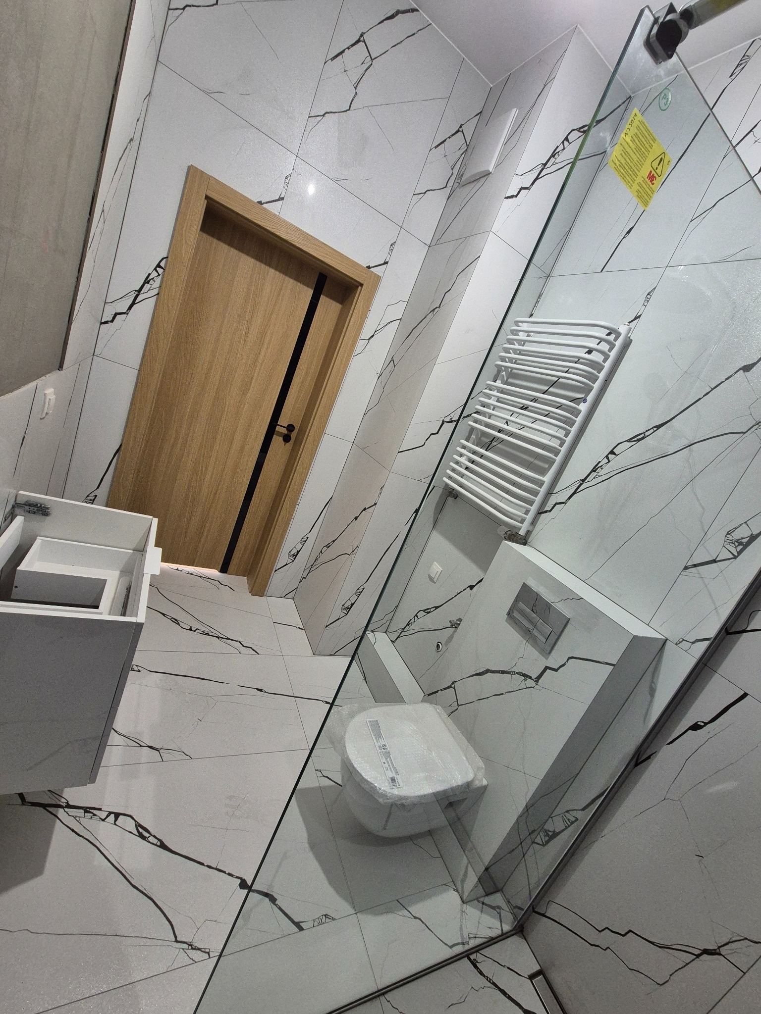 Nowoczesna łazienka z białymi płytkami imitującymi marmur, kabiną prysznicową ze szkła, grzejnikiem drabinkowym i drewnianymi drzwiami z czarnymi akcentami.