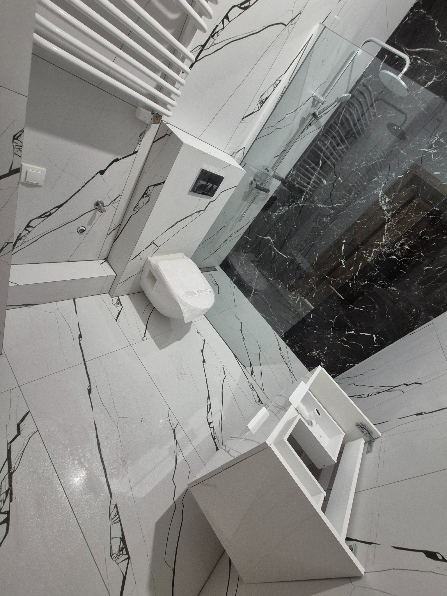 Nowoczesna łazienka z kabiną prysznicową i umywalką w Grodzisku Mazowieckim. Białe płytki imitujące marmur z czarnymi żyłami na ścianach i podłodze.