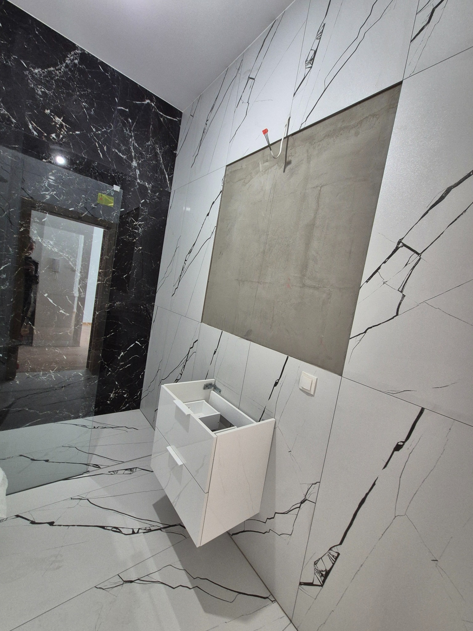 Łazienka w trakcie wykończenia: białe i czarne płytki imitujące marmur, zamontowana szafka pod umywalkę, fragment ściany bez płytek przygotowany pod lustro.