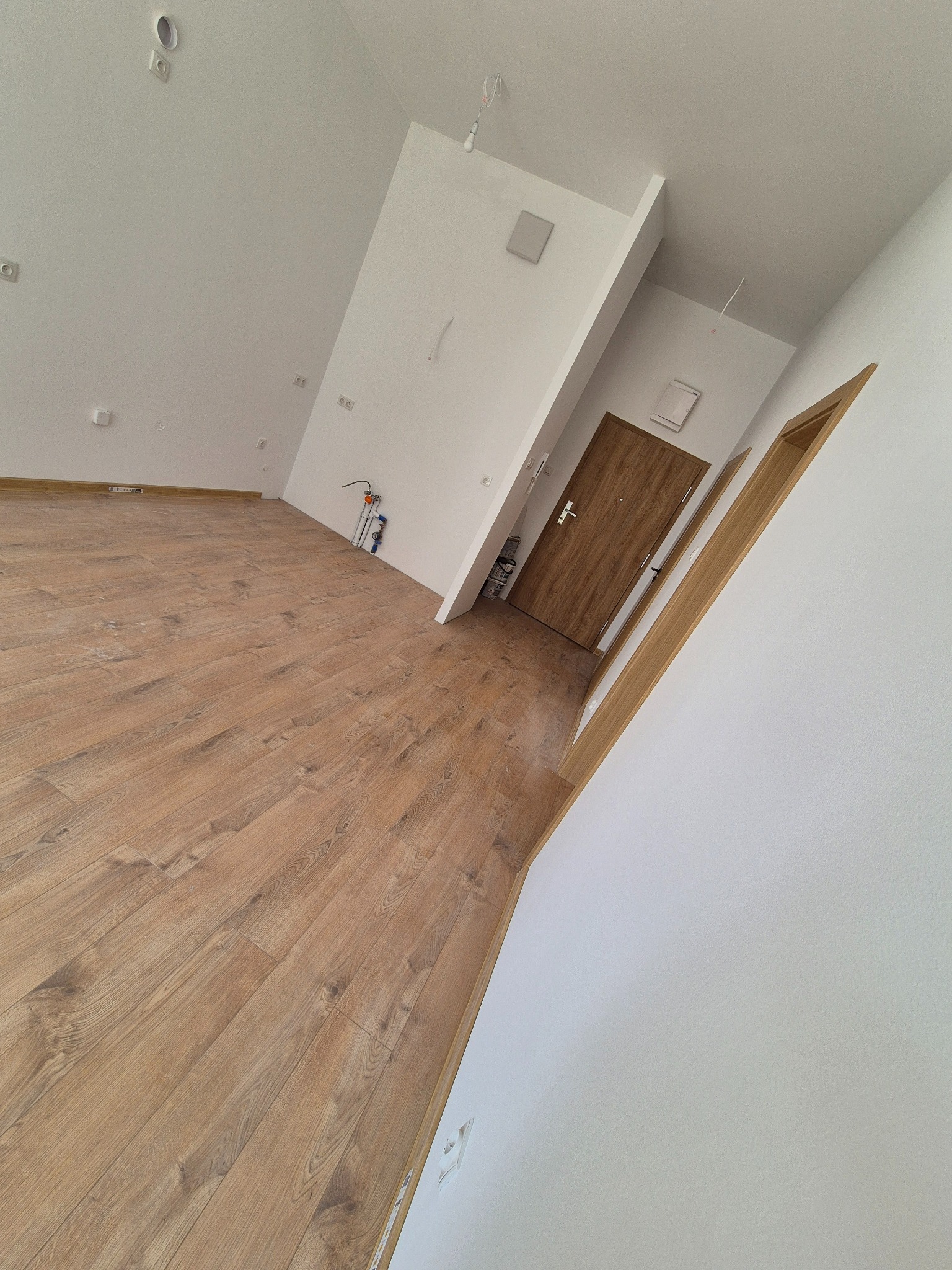 Jasne wnętrze po remoncie: drewnopodobne panele, białe ściany, drzwi wejściowe z brązowego drewna. Widoczne gniazdka i elementy instalacji.