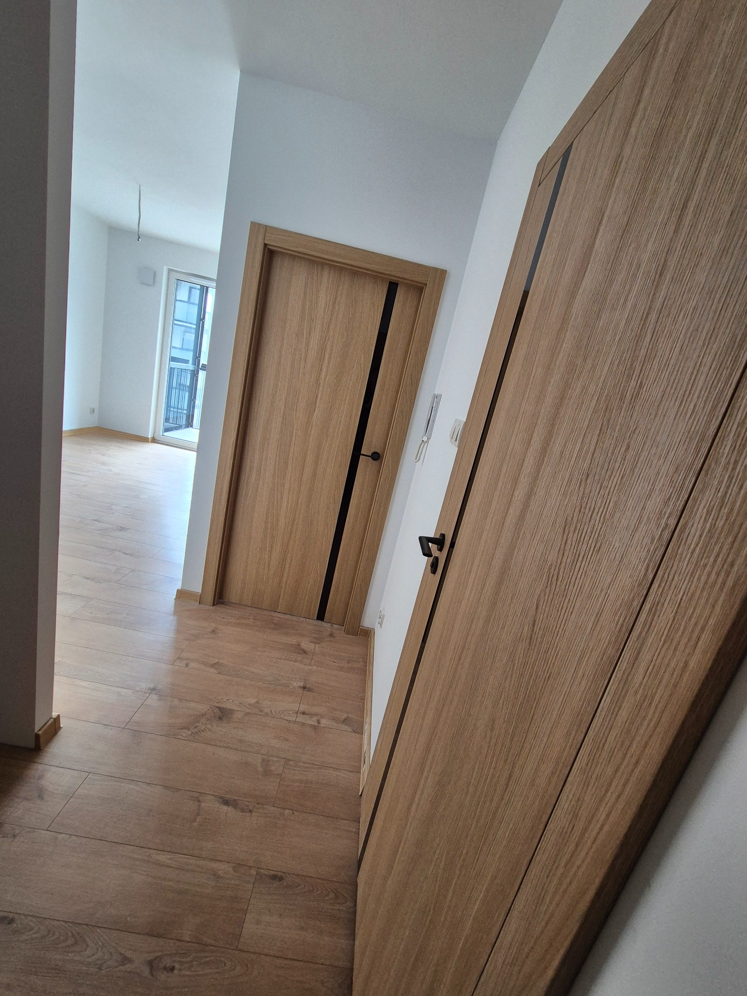 Nowe drewniane drzwi wewnętrzne z czarnymi akcentami, widok na parkiet i odległe okno w wyremontowanym mieszkaniu. Minimalistyczny design.