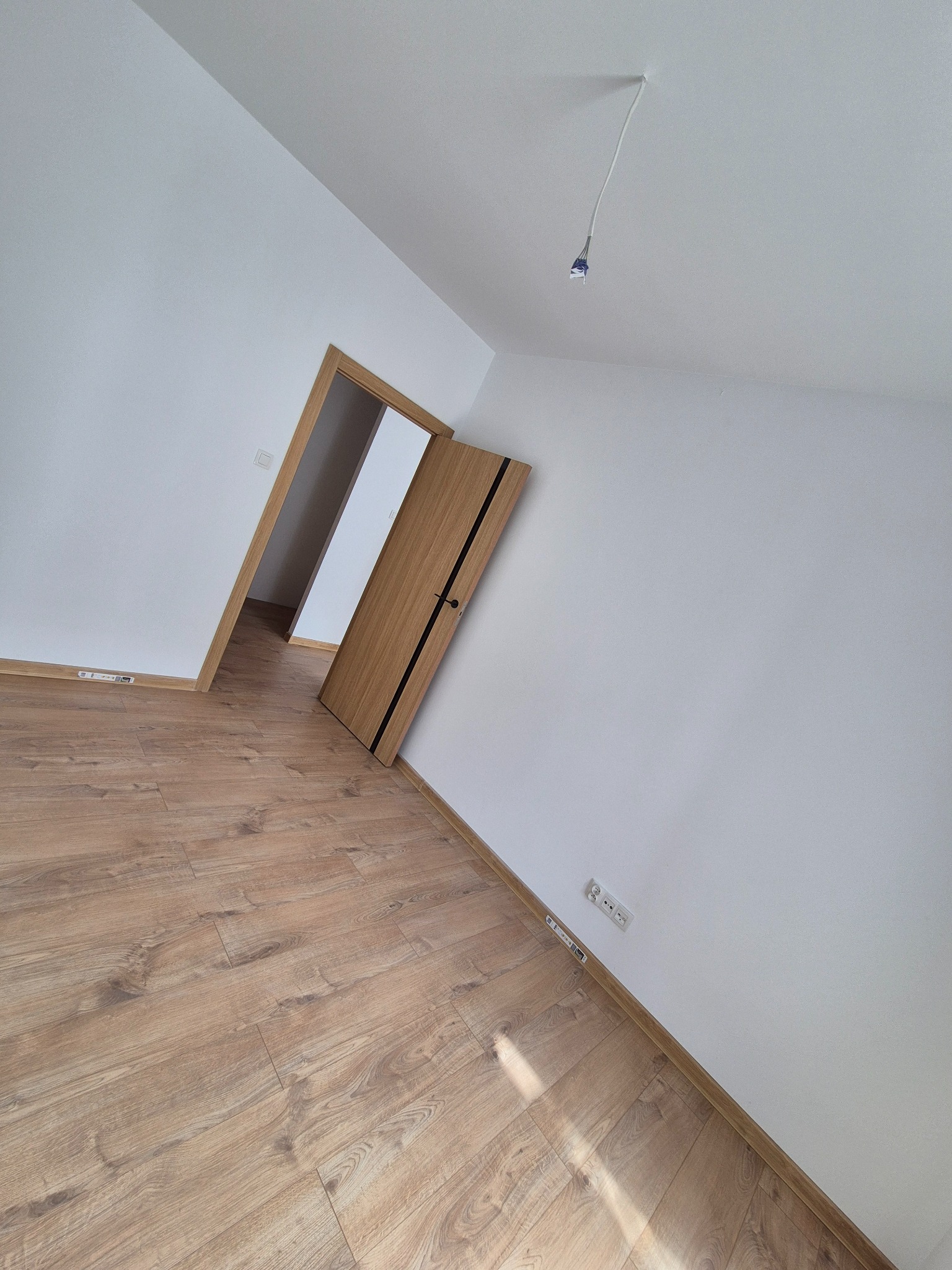 Jasne panele podłogowe w pokoju z białymi ścianami i drewnianymi drzwiami z czarnymi akcentami. Widoczne gniazdka elektryczne i wystający kabel z sufitu.