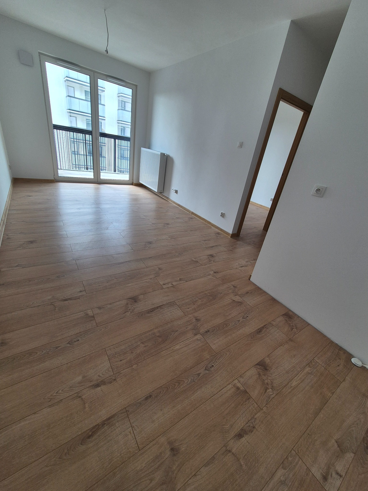 Jasne panele podłogowe w nowym, pustym pokoju z balkonem i grzejnikiem. Widok na otwarte drzwi do sąsiedniego pomieszczenia. Minimalistyczny design.