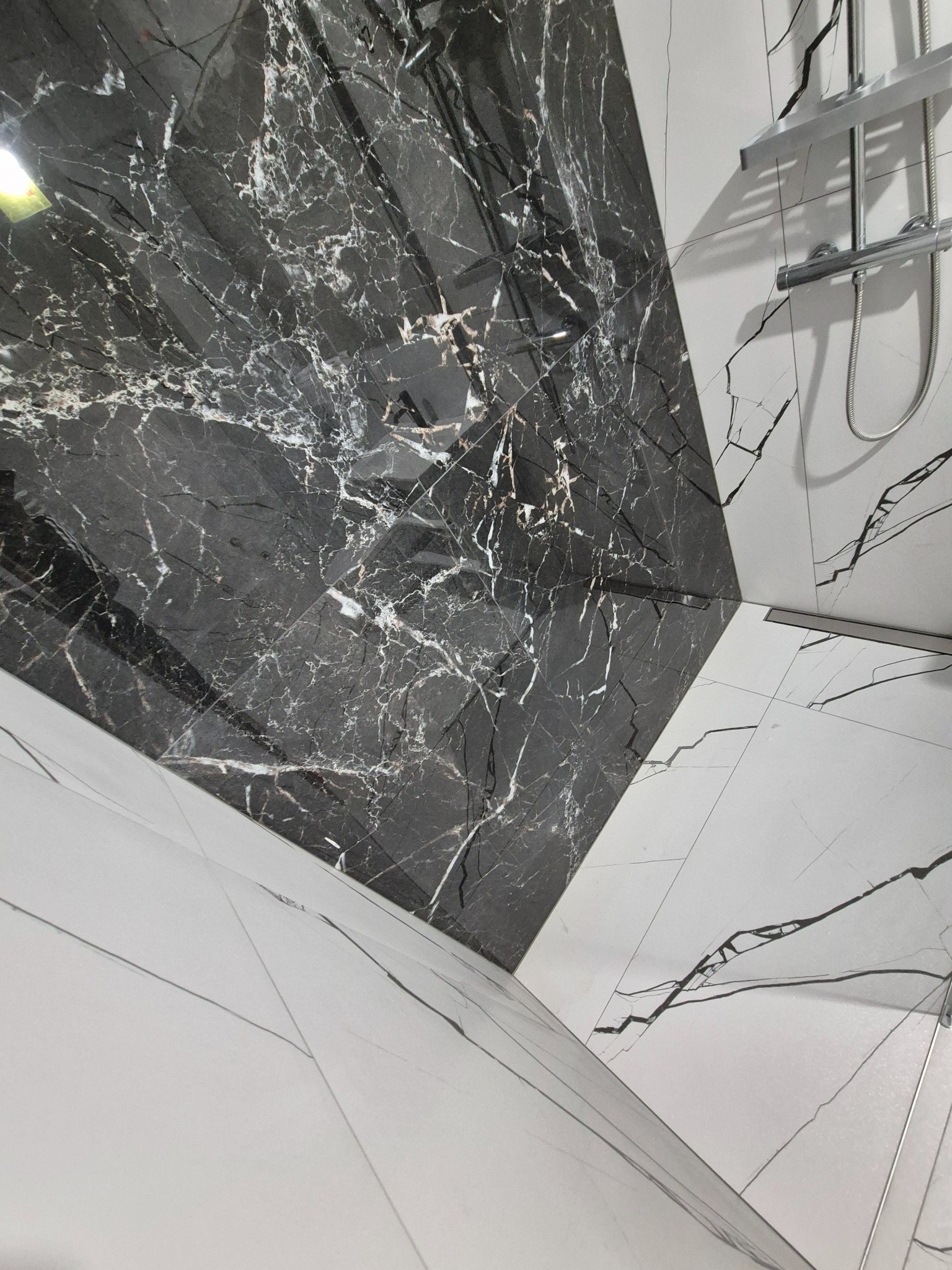 Nowoczesna łazienka z czarno-białymi płytkami imitującymi marmur ułożonymi pod kątem, widoczna armatura prysznicowa i odpływ liniowy.