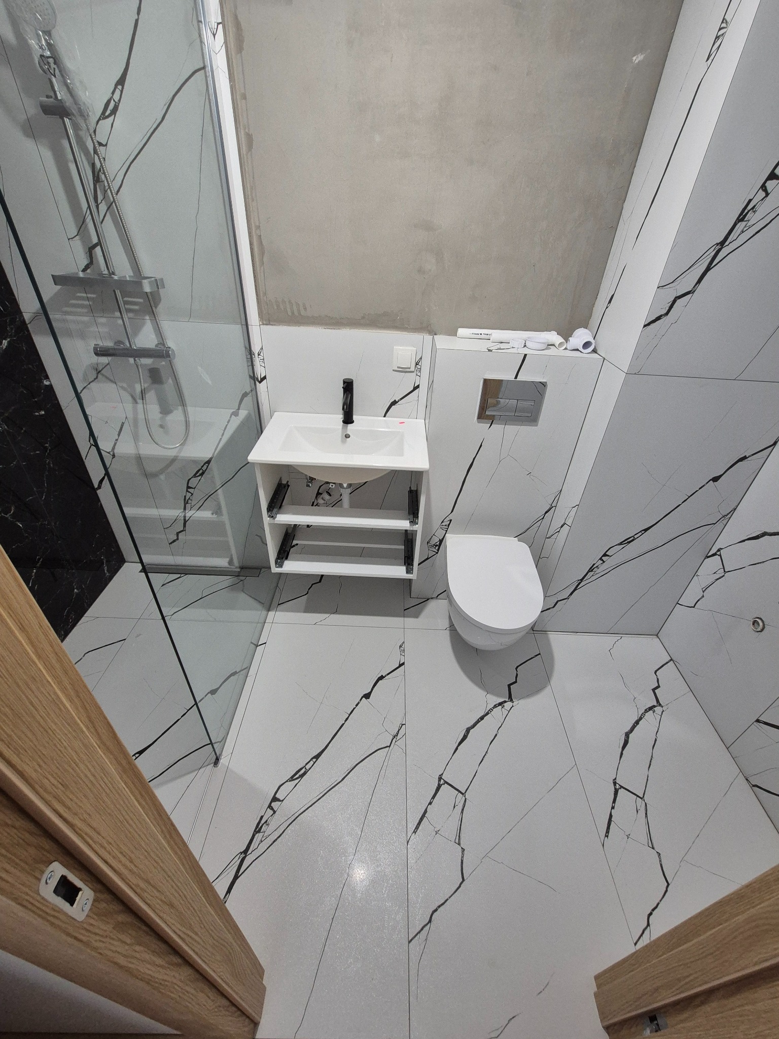 Biała łazienka w stylu marmuru z kabiną prysznicową, umywalką z szafką i toaletą. Surowy, betonowy sufit kontrastuje z eleganckimi płytkami.