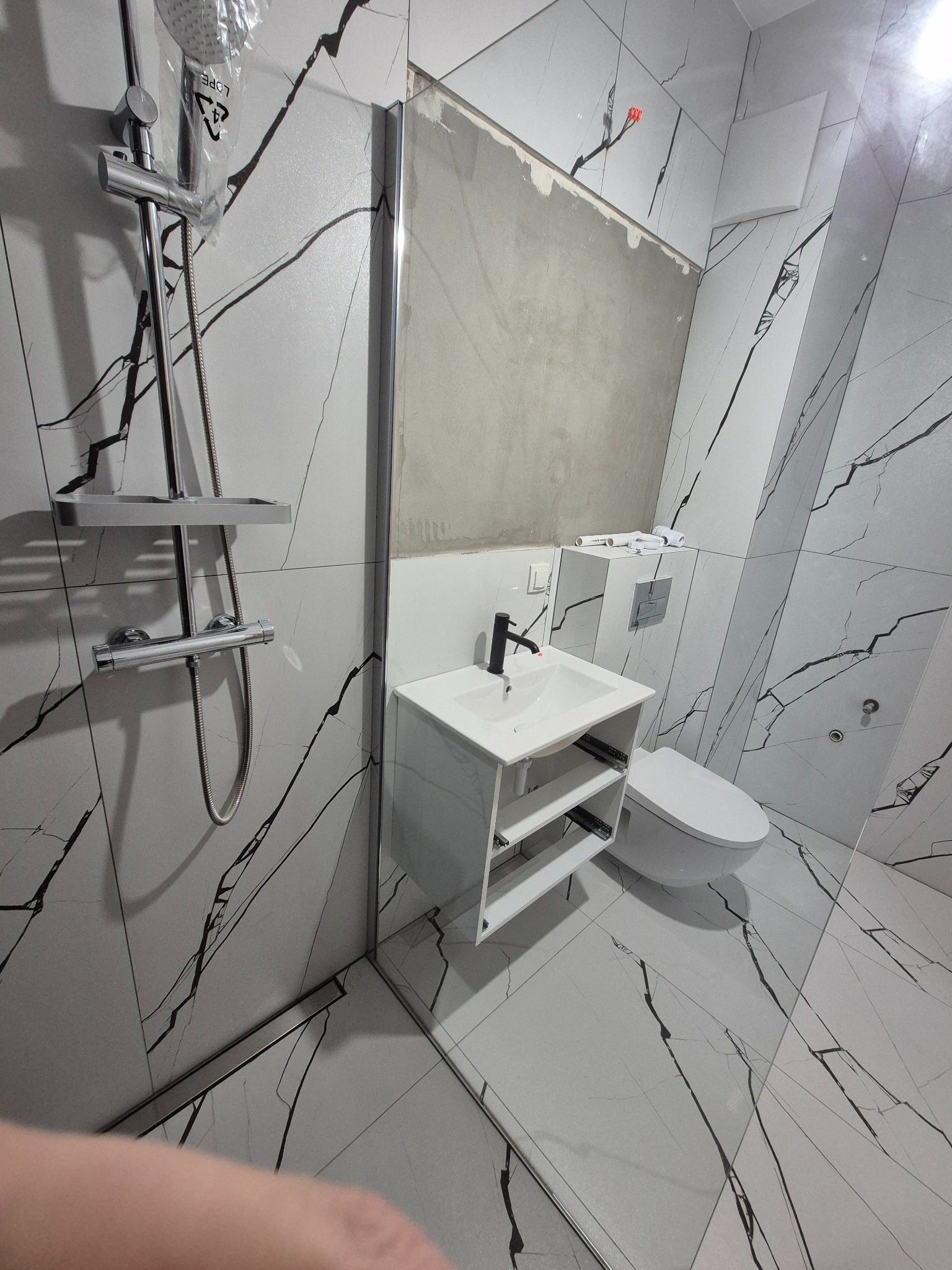 Nowoczesna łazienka w trakcie wykańczania: białe płytki z czarnym wzorem, umywalka z szafką i toaleta. Widoczny niedokończony montaż lustra.