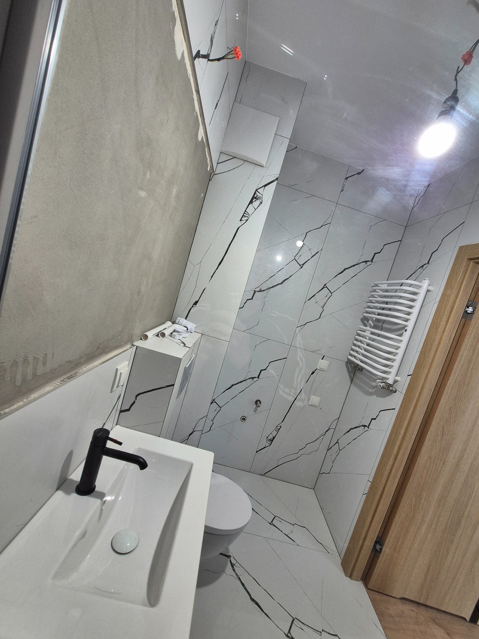 Wnętrze łazienki w trakcie remontu: białe płytki z czarnym wzorem marmuru, umywalka z czarną armaturą, grzejnik drabinkowy. Częściowo wykończona ściana z gładzią.